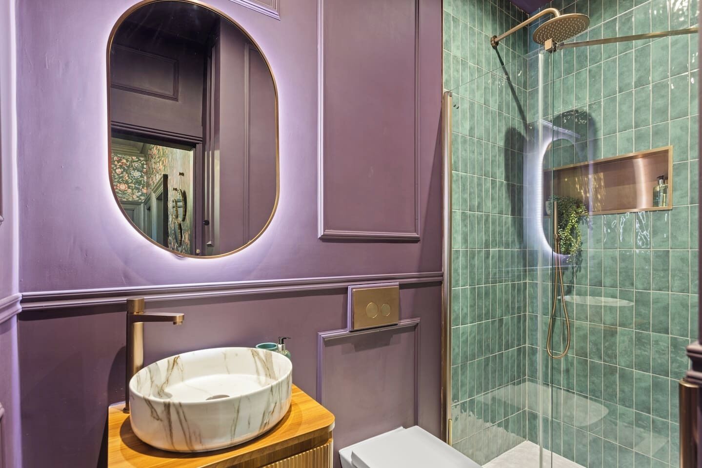 A bathroom with a marble sink, a round mirror, a shower with a glass door, and a green tiled wall.
