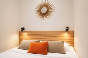 Cama com cabeceira de madeira, duas luminárias fixadas na parede e espelho em forma de sol.