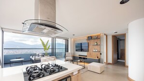 Een moderne keuken met een gasfornuis, een groot raam met uitzicht op een stadsgezicht, en een zithoek met een televisie en houten planken.