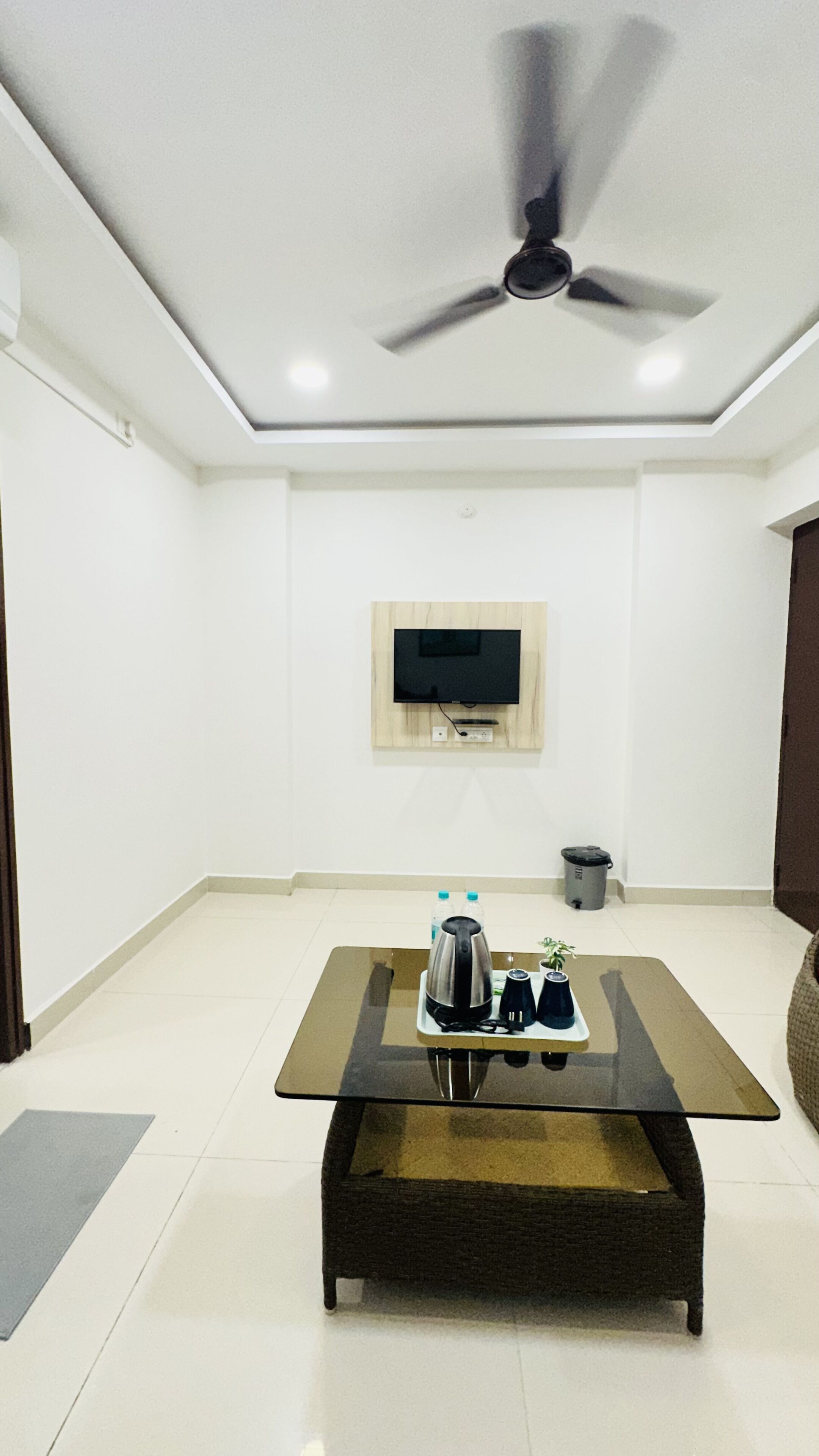 A modern living room with a glass-top coffee table, a ceiling fan, a mounted TV, and a trash bin.