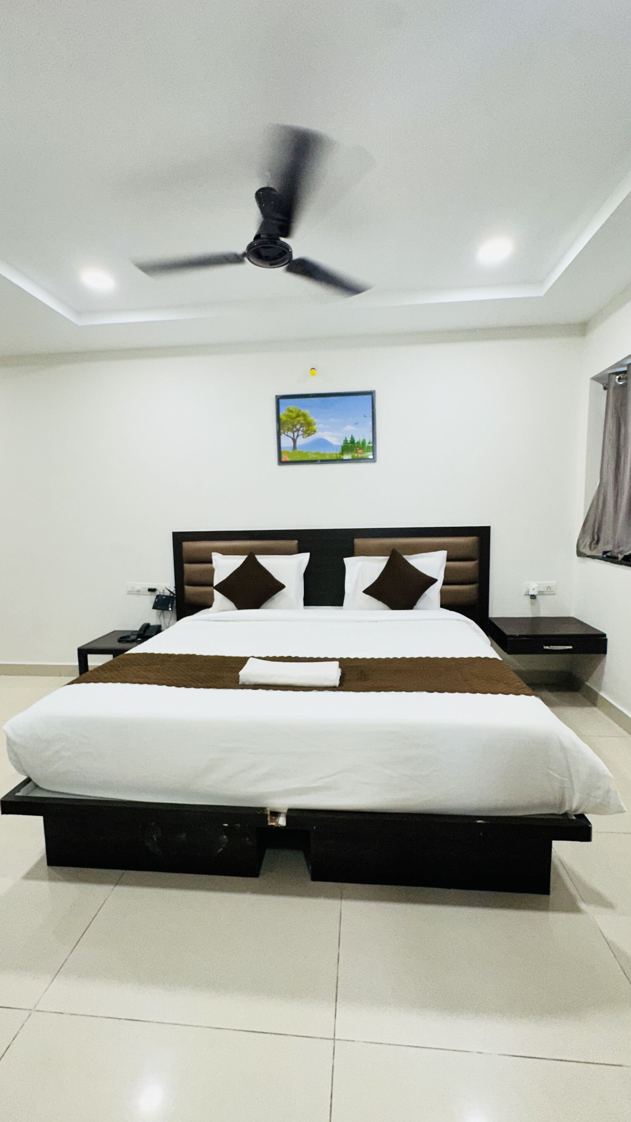 A modern hotel room with a bed, a ceiling fan, a nightstand, a phone, and a picture on the wall.