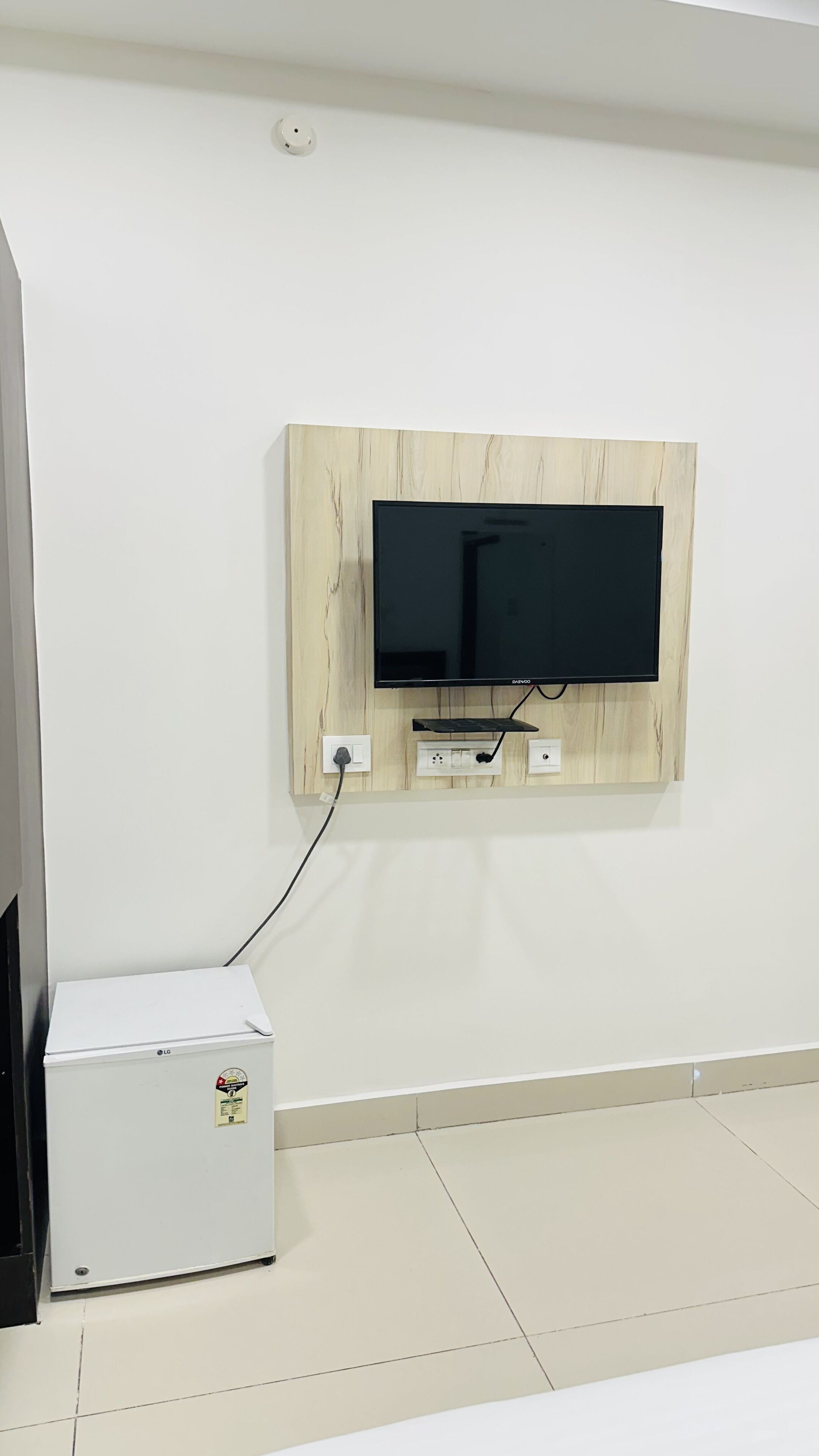 A flat-screen TV mounted on a light wood panel wall, connected to a white LG refrigerator on the floor.