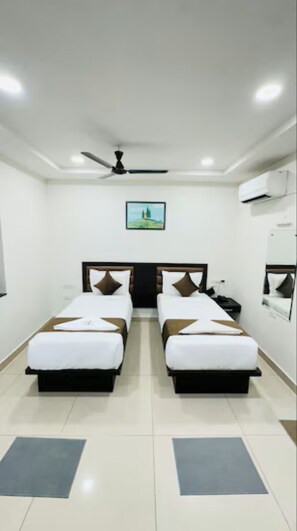 A hotel room with two beds, a ceiling fan, an air conditioning unit, a framed picture on the wall, and a small table with a chair. - Innzoy Comforts (Hyderabad)