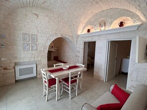 A dining area with a table and chairs, a kitchen visible through an archway, and a television on the wall. - Complex of trulli and lamie in Itria Valley - charm and relaxation by the pool (Puglia)