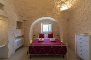 A bedroom with a stone wall, a bed with a red bedspread, a window, and a white dresser. - Complex of trulli and lamie in Itria Valley - charm and relaxation by the pool (Puglia)