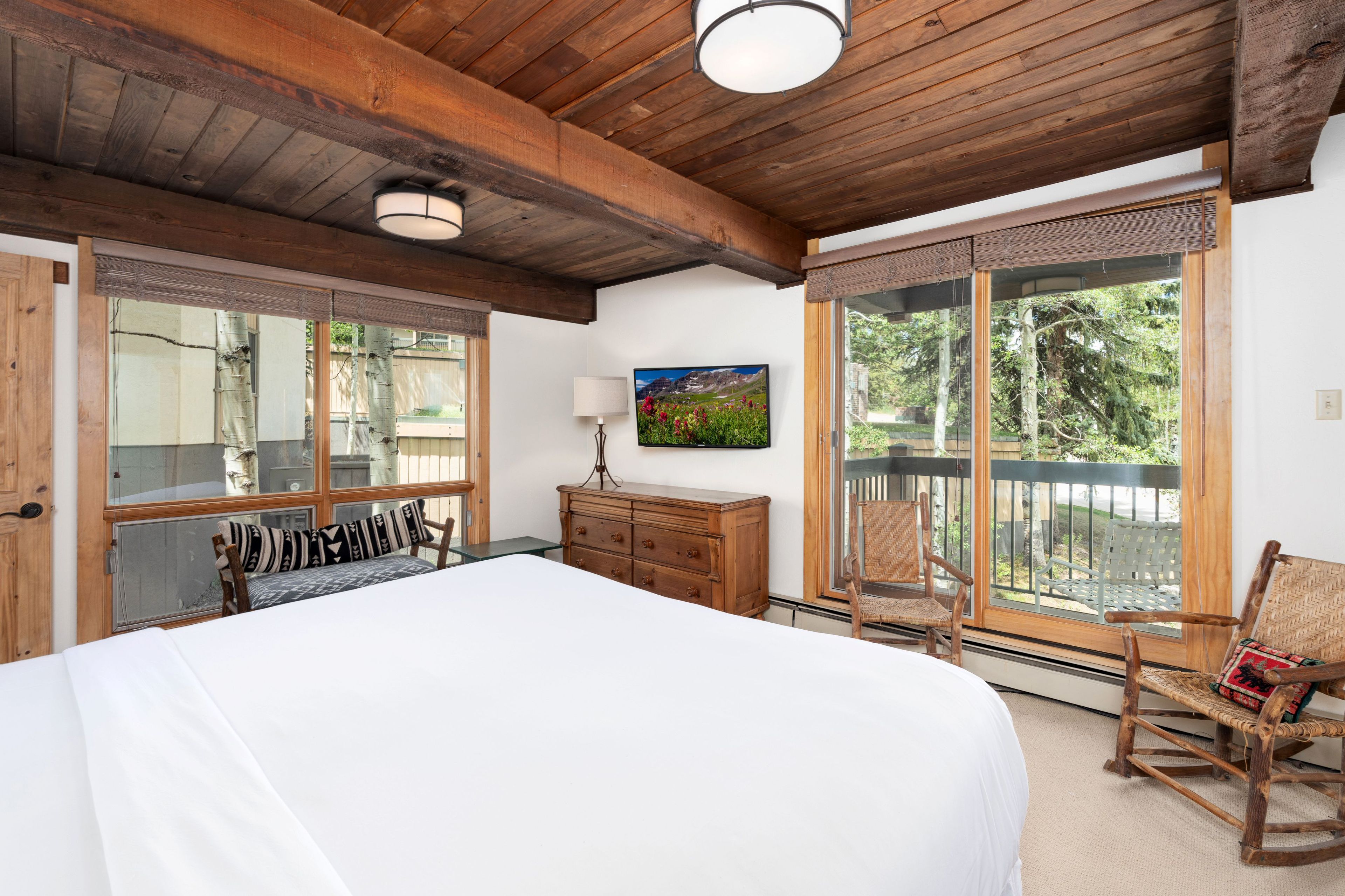 Une chambre avec un grand lit, un plafond en bois, une commode, une chaise et une fenêtre donnant sur les arbres.