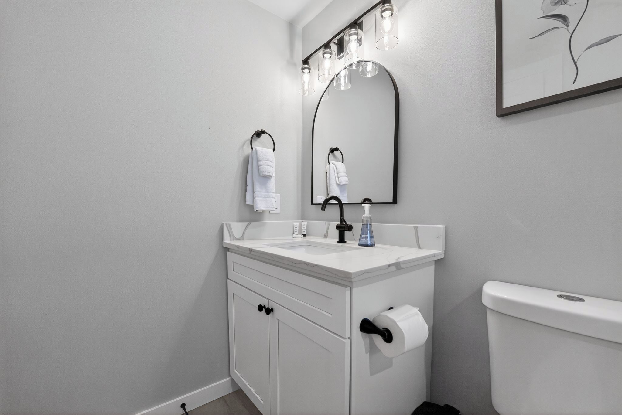 A bathroom with a white sink, black faucet, and a toilet. There is a mirror with lights above it and a towel hanging on a hook.