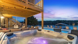 A hot tub with lounge chairs, a wooden deck, and a view of a swimming pool and trees at dusk. - Lakefront Sleeps 18 w/ Pool, Dock, Putting Green, Hot Tub! Hot Springs (Hot Springs)