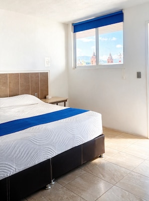 Habitación de hotel con cama, mesita de noche, ventana con vista a edificios y suelo de baldosas.