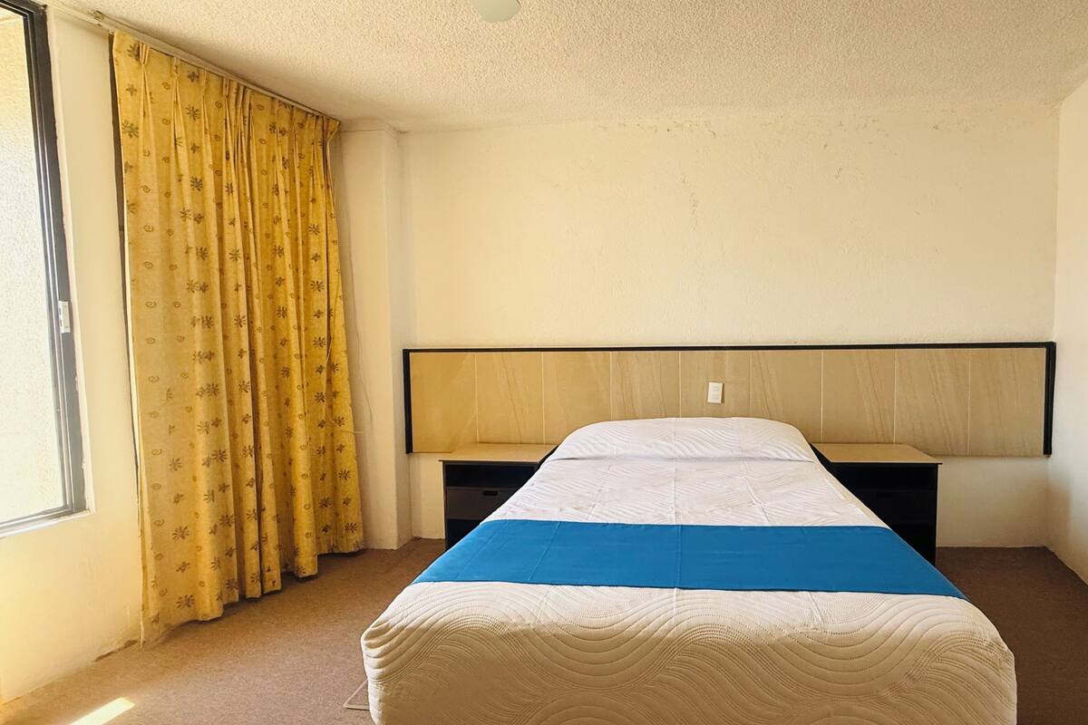 Una habitación de hotel con cama, mesita de noche, ventana con cortinas y techo sencillo.