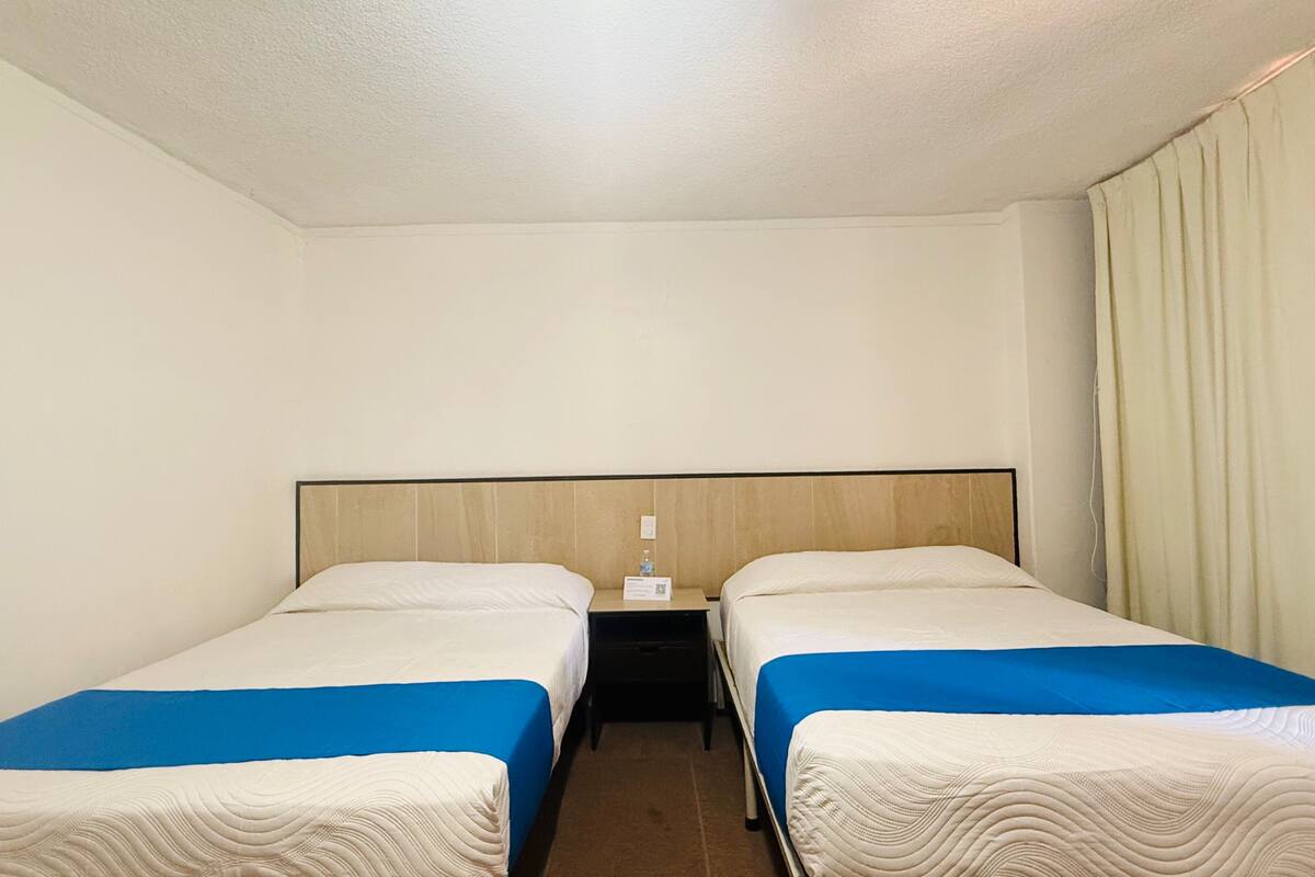 Una habitación de hotel con dos camas individuales, cabecera de madera, mesita de noche con lámpara y cortina en la ventana.