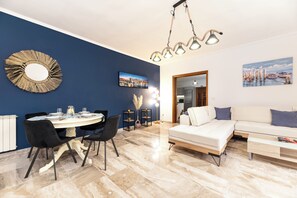 A modern living room with a sofa, dining table, and chairs. There is a painting on the wall and a lamp.