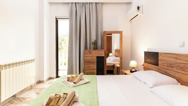 Quarto de hotel com cama, mesa de cabeceira, espelho, janela com cortinas e radiador.