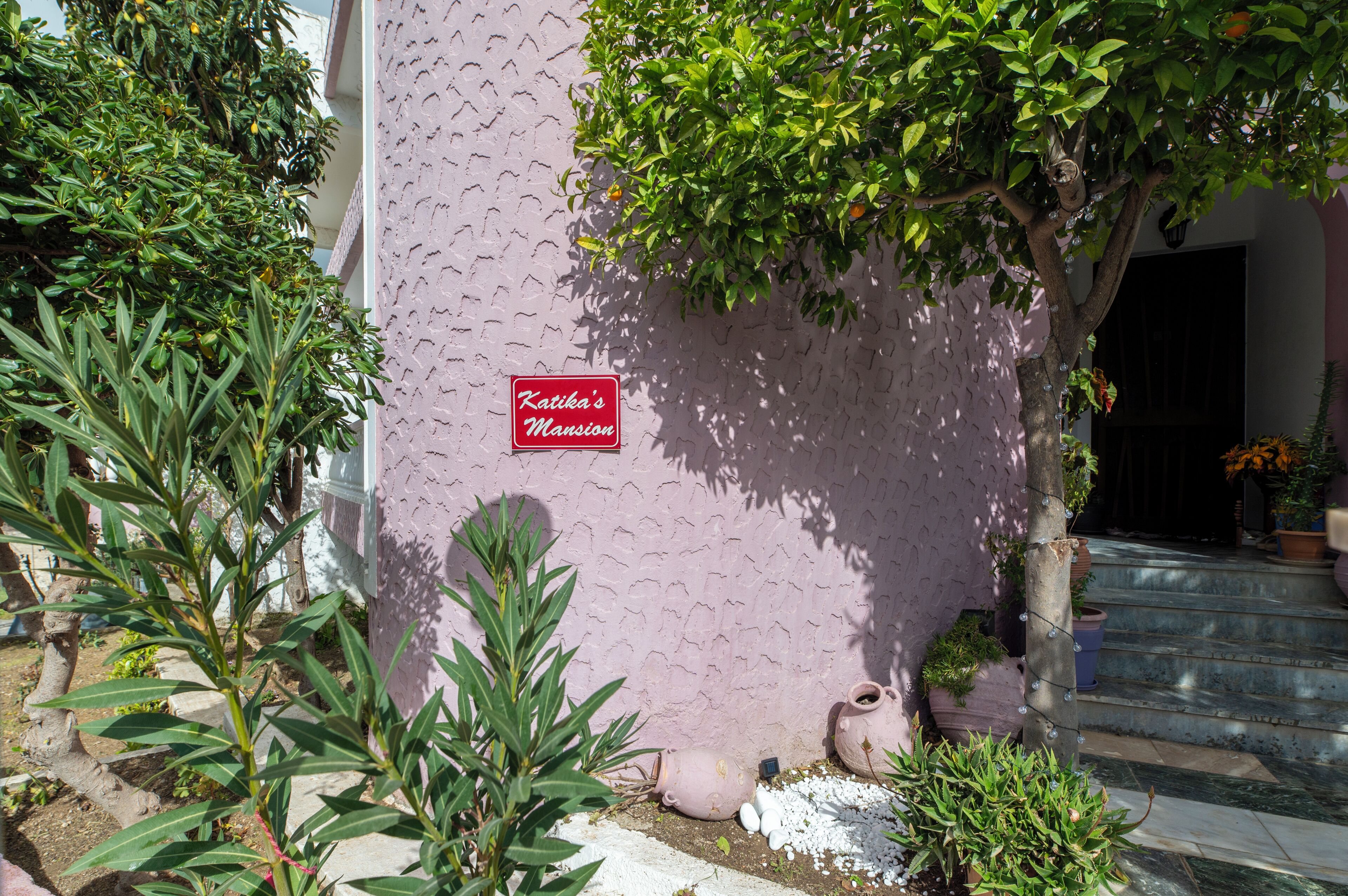 A pink building with a sign reading 'Katika's Mansion,' surrounded by greenery and potted plants.
