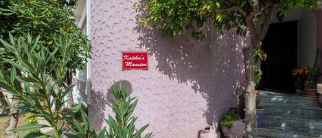 A pink building with a sign reading 'Katika's Mansion,' surrounded by greenery and potted plants.