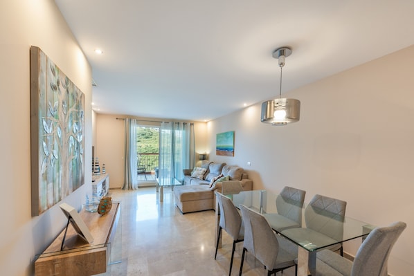 A modern living room with a glass dining table, a sofa, and a large abstract painting. - Alcaidesa Golf and Beach Resort: Relaxation with Pool, Mountain Views and Prime Location (La Alcaidesa)