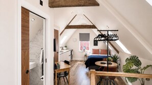 Ein Loft-Zimmer mit schrägem Dach, Holzbalken, einem Essbereich, einer Küche und einem großen Fenster.