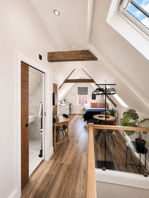 Ein Loft-Zimmer mit schrägem Dach, Holzbalken, einem Essbereich, einer Küche und einem großen Fenster.
