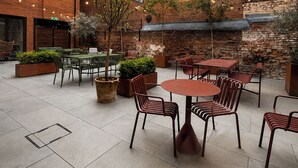 Eine Terrassengaststätte mit Tischen und Stühlen, umgeben von Backsteingebäuden und Grünpflanzen.