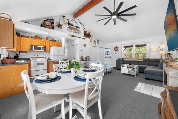 Une cuisine moderne avec des meubles blancs, une table à manger avec des chaises, un canapé gris et un ventilateur de plafond.