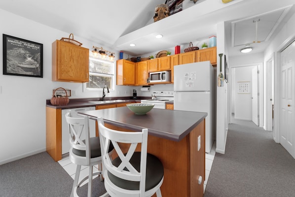 A modern kitchen with wooden cabinets, a central island, white chairs, and a refrigerator.