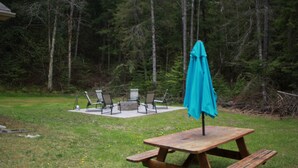 Ett picknickbord i trä med bänkar, ett turkosfärgat parasoll och utomhusstolar på en gräsig bakgård med träd i bakgrunden.