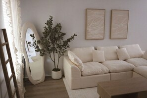 A living room with a white sofa, a wooden coffee table, a potted plant, a mirror, and two framed artworks on the wall.