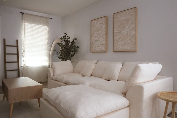 Un canapé blanc, une petite table en bois et deux œuvres d’art encadrées accrochées au mur, dans une pièce où se trouvent une échelle et une plante.