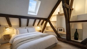 Ein Schlafzimmer mit schrägverdeckter Decke, freiliegenden Holzbalken, einem Bett mit weißer Bettwäsche, Nachttischlampen und einem kleinen Tisch mit einer Lampe.