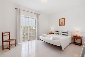 Ein Schlafzimmer mit Bett, Sessel, Nachttisch, einem Bild an der Wand und einer Schiebetür zum Balkon.