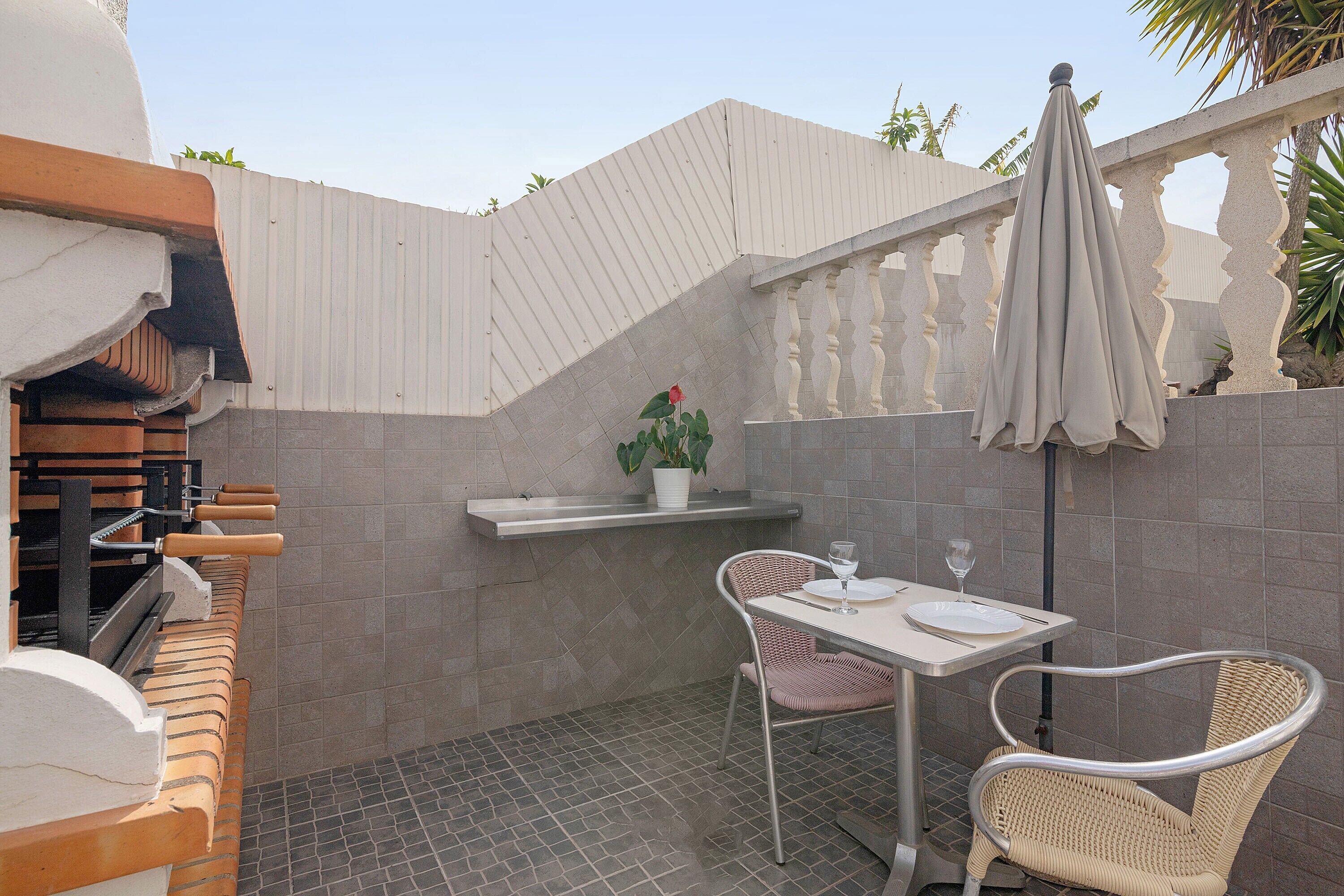 Un patio con una parrilla, una mesa con sillas, una planta en maceta y un paraguas decorativo.