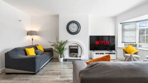 A modern living room with a sofa, a TV, a fireplace, and a clock on the wall. - 6-bedroom home for contractors and families. (Colchester)