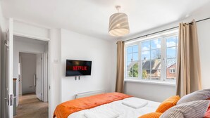 A bedroom with a bed, a TV mounted on the wall, a window with curtains, and a hallway leading to another room. - 6-bedroom home for contractors and families. (Colchester)