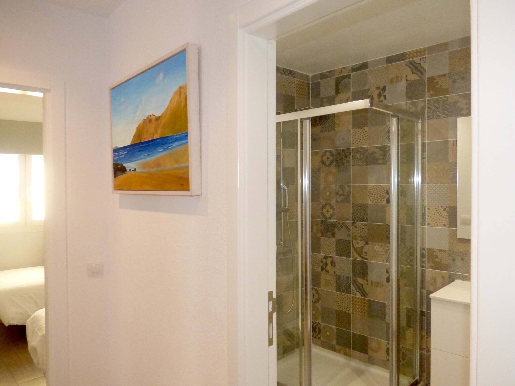 Un baño con baldosas estampadas, mampara de vidrio y un cuadro con una escena de playa en la pared.
