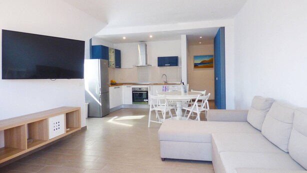 Uma sala de estar moderna com um sofá branco, uma TV na parede e uma área de cozinha com uma mesa de jantar e cadeiras.