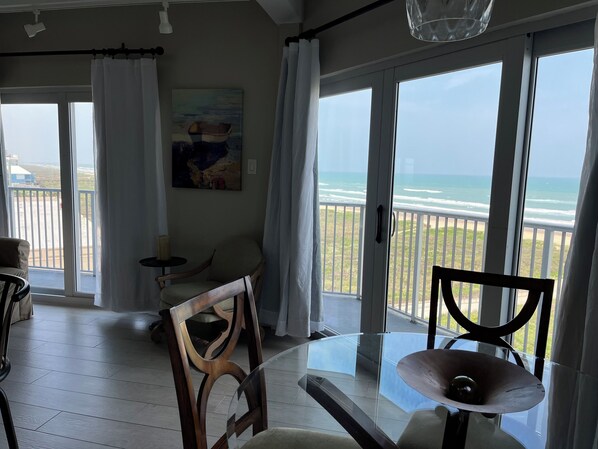 Ein Zimmer mit Glastisch, Holzstuhl, einem Gemälde an der Wand und Blick auf den Strand durch große Fenster.