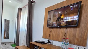 A modern hotel room with a wooden desk, a grey chair, a flat-screen TV mounted on the wall, and a small shelf with a kettle and a red tray.