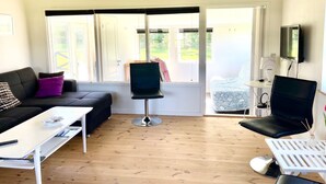 Un salon moderne avec un canapé, une table basse et deux fauteuils. Un téléviseur est fixé au mur et il y a une fenêtre avec des stores.