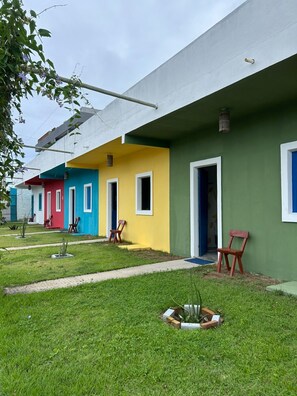 A row of colorful buildings with doors and windows, a grassy area with chairs, and a small garden bed. - Pousada Villa Japa (Maragogi)