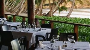 An outdoor dining area with tables set for two, overlooking a beach with palm trees. - La Sagesse Bay View (St. David)