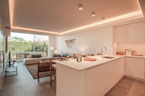 A modern kitchen with a central island, a dining area with a sofa, and a large window providing a view of the outdoors. - FLH Vilamoura Boho Apartment with Terrace (Vilamoura)