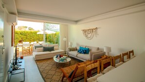 A modern living room with a sofa, coffee table, and chairs, a dining area with a table and chairs, and a large window with a view of greenery. - FLH Vilamoura Boho Apartment with Terrace (Vilamoura)