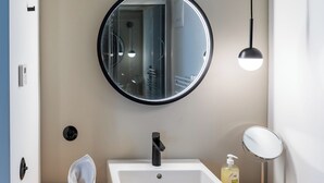 Ein modernes Badezimmer mit einem runden Spiegel, einem weißen Waschbecken, einem schwarzen Wasserhahn, einem kleinen Seifenspender und einem Handtuch auf der Ablagefläche.