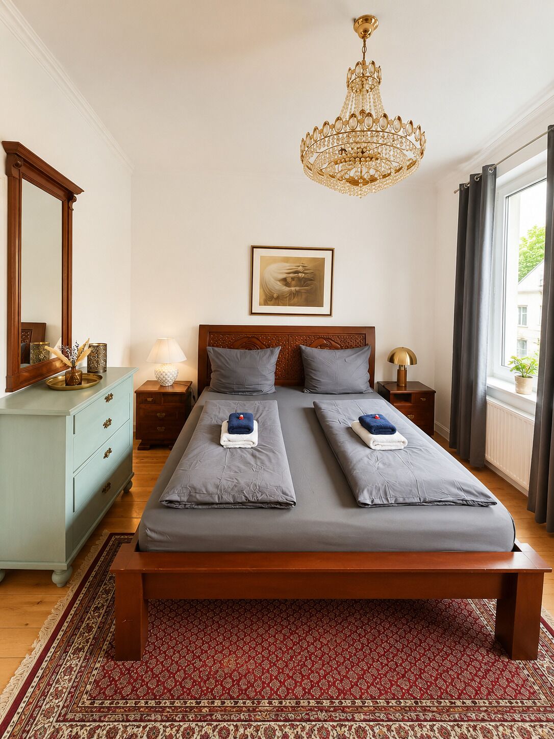 A bedroom with a large bed, a chandelier, a bedside table, a dresser, a mirror, and a window with curtains.