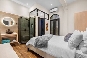 Ein modernes Schlafzimmer mit Bett, einer Duschkabine aus Glas, einem Holzwaschtisch mit Schüsselspüle und einer wandmontierten Klimaanlage.