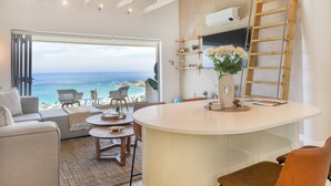 Ein modernes Wohnzimmer mit großem Fenster zum Meer, einem weißen runden Tisch und einer eingebauten Leiter.