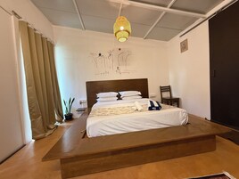 Un dormitorio con una cama, una plataforma de madera, una lámpara colgante, una silla y una pared con un dibujo sencillo.