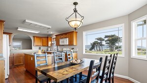 A kitchen with wooden cabinets, a dining table with chairs, a chandelier, and a view of trees through large windows. - Beach House | Oceanfront + Fenced Yard (Crescent City)