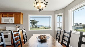A dining area with a wooden table, chairs, a chandelier, and a view of a landscape through large windows. - Beach House | Oceanfront + Fenced Yard (Crescent City)