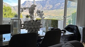 Un salon moderne avec vue sur les montagnes, une grande fenêtre et une table ornée d’un vase de fleurs blanches.